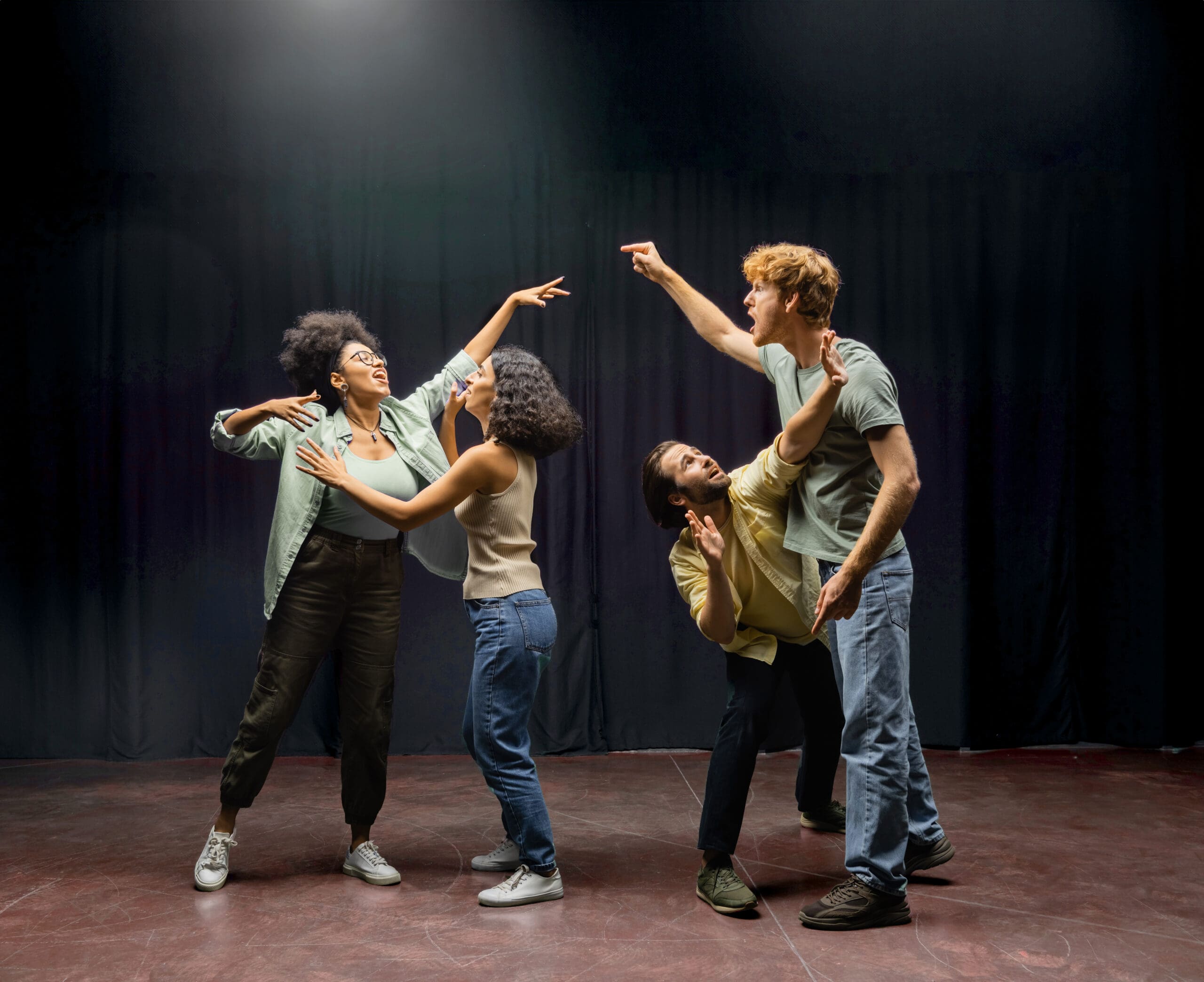 Quatre personnes sont dans une pose dramatique sur une scène faiblement éclairée. Deux d'entre elles sont debout avec des expressions animées utilisant des gestes expressifs des mains, tandis que les deux autres sont à genoux, en train de dialoguer intensément. L'arrière-plan est sombre avec un projecteur au-dessus.